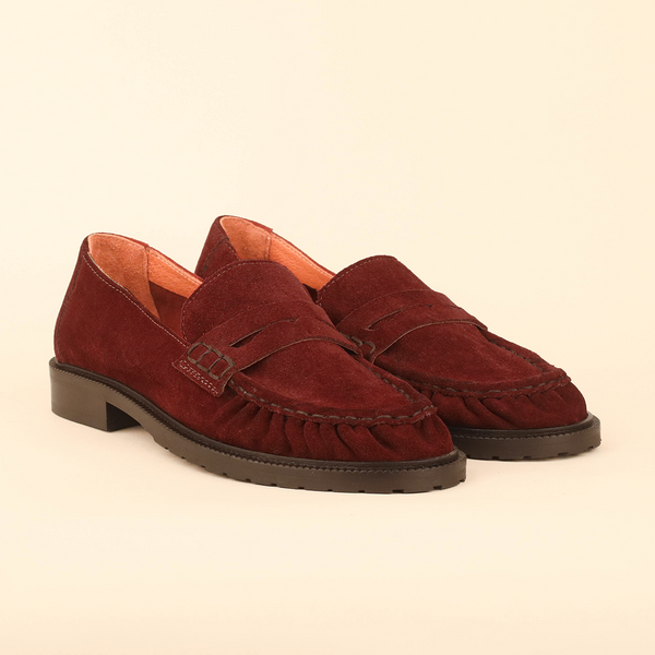 A pair of rust red suede penny loafers with dark brown soles, displayed on a warm cream background.