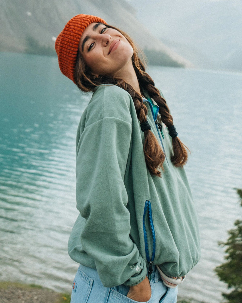 Model stands by a lake and wears sage green recycled polyester fleece and orange beanie hat.