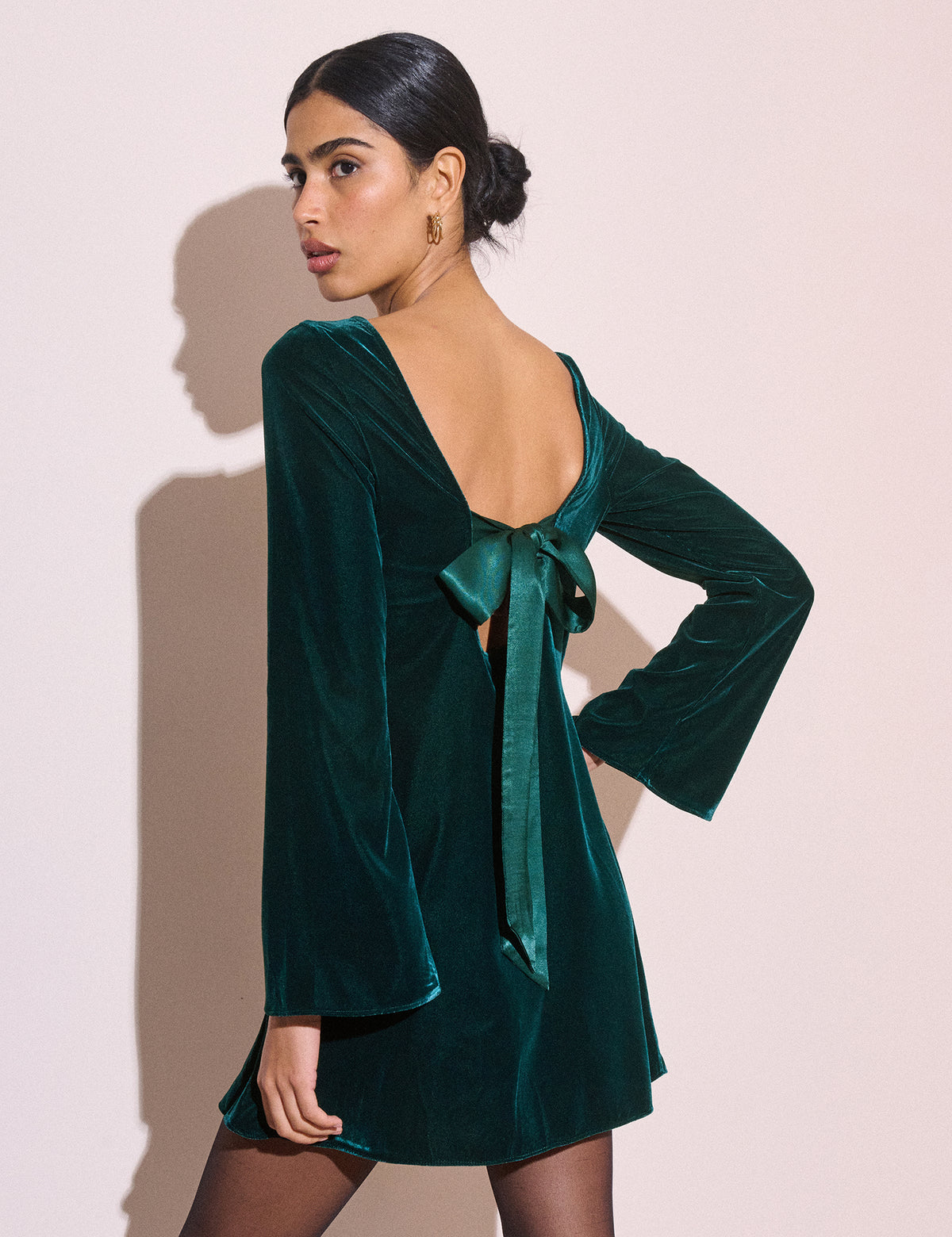 A woman modeling a dark teal velvet mini dress with long bell sleeves, a deep open back, and a matching bow tie at the waist, posed against a solid pale pink background.