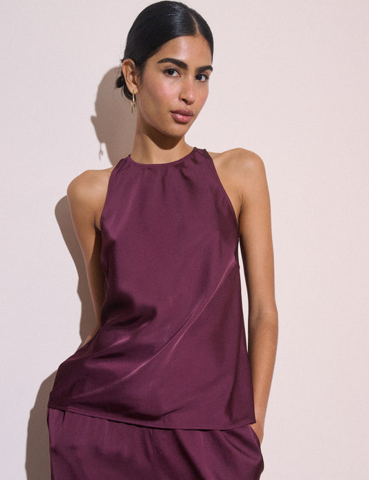A woman modeling a sleeveless satin-like top in a deep plum purple color, paired with matching bottoms, against a plain light beige wall.