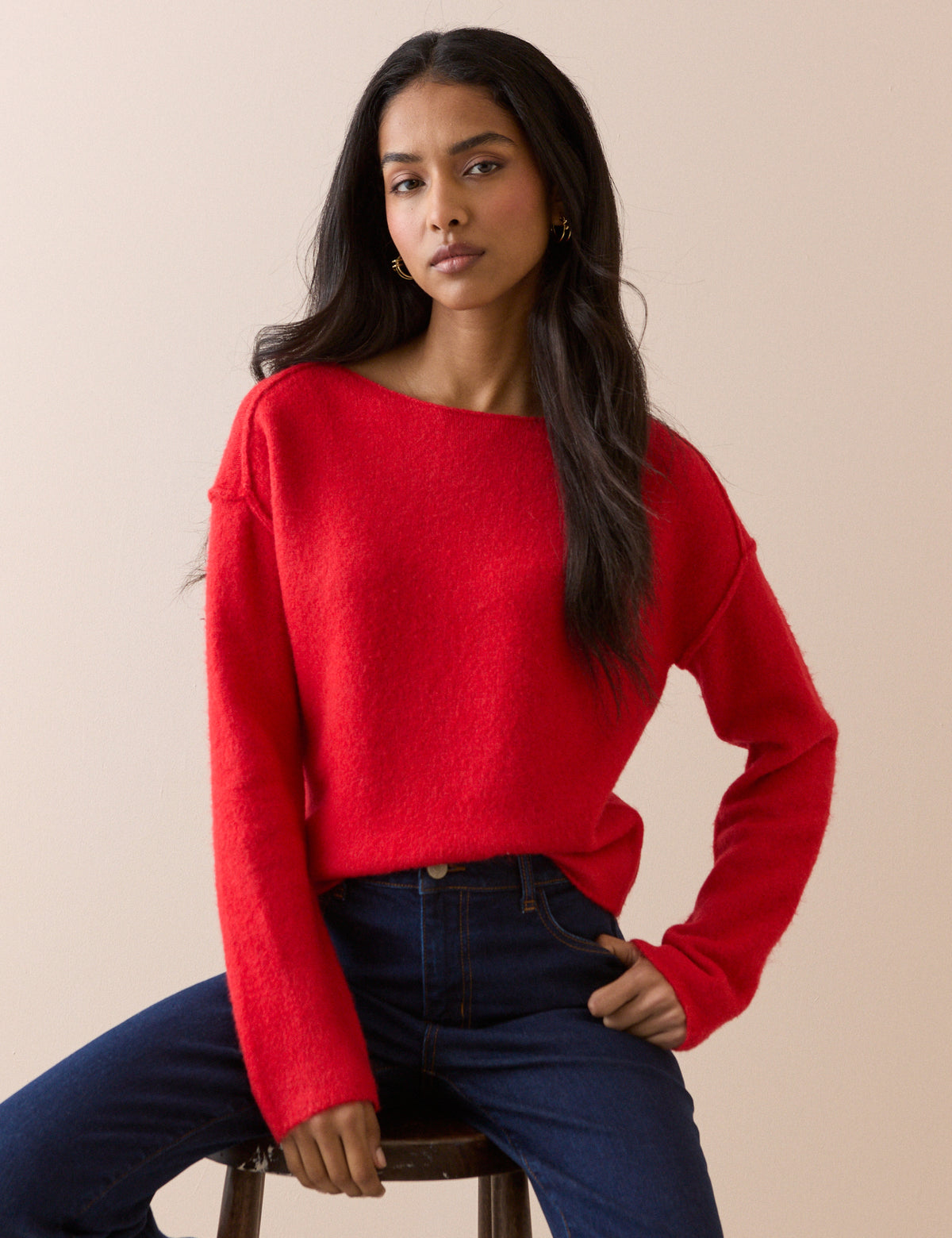A woman wearing a bright red knit jumper and dark blue jeans, seated on a wooden stool against a solid beige background.