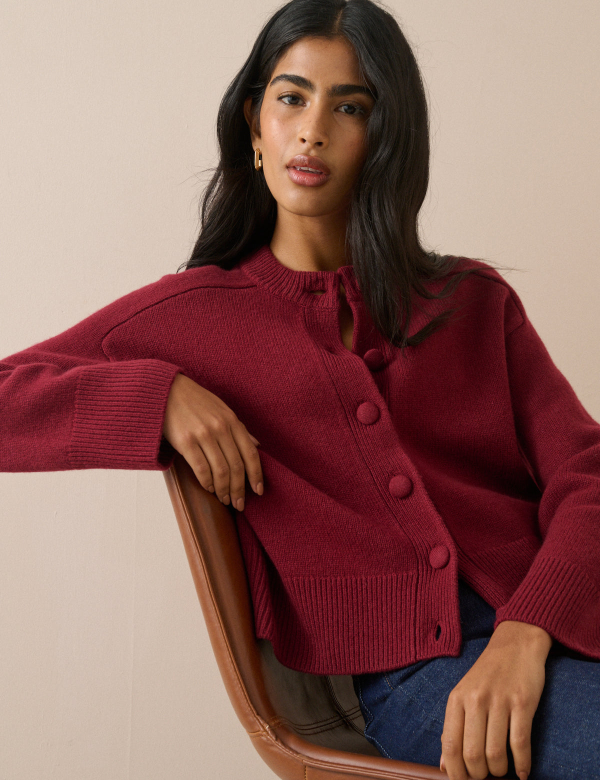 A woman wearing a burgundy knit cardigan sweater with a tie-neck detail and large front buttons, seated on a brown leather chair against a plain beige background.