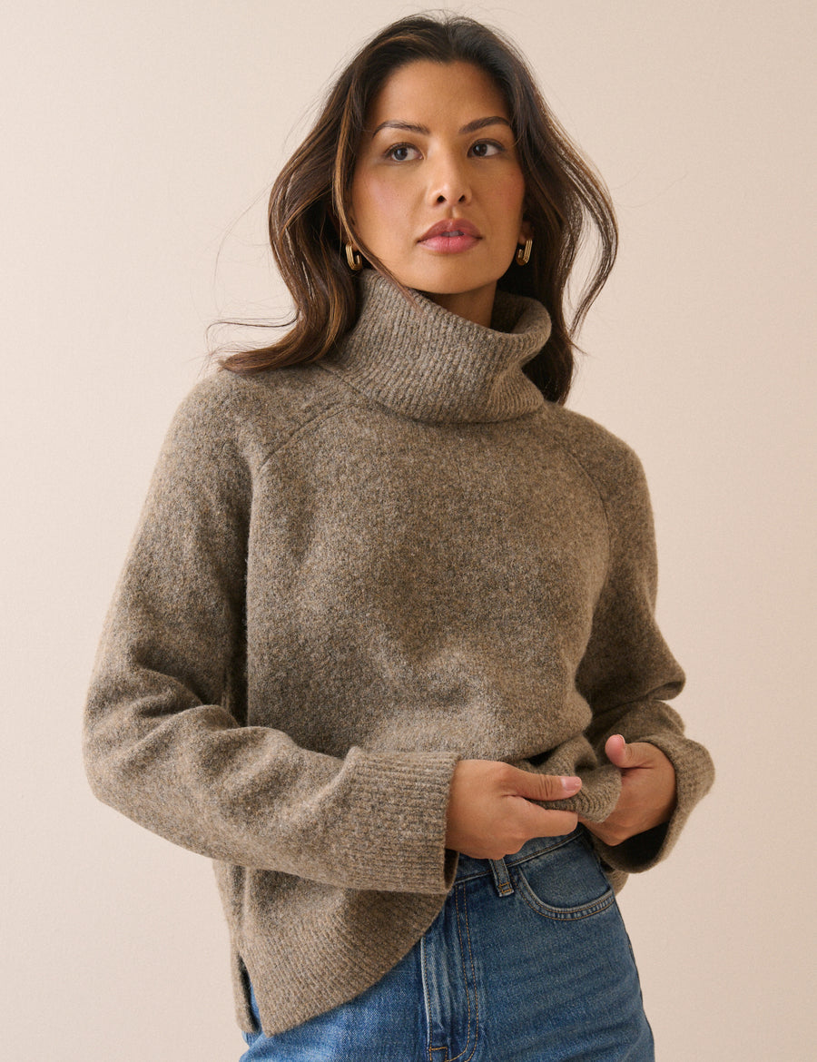 A woman wearing a fuzzy, heathered taupe-brown roll neck pullover and blue jeans, posing against a solid light beige background.