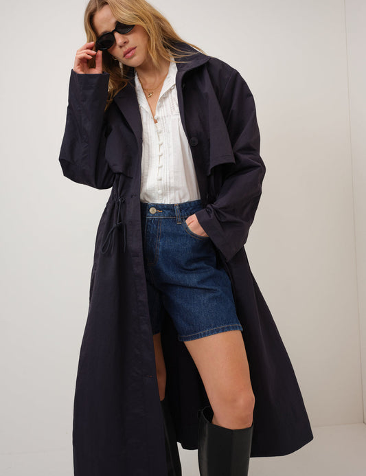Woman wearing a long dark coat over a white shirt and denim shorts, standing against a plain background.
