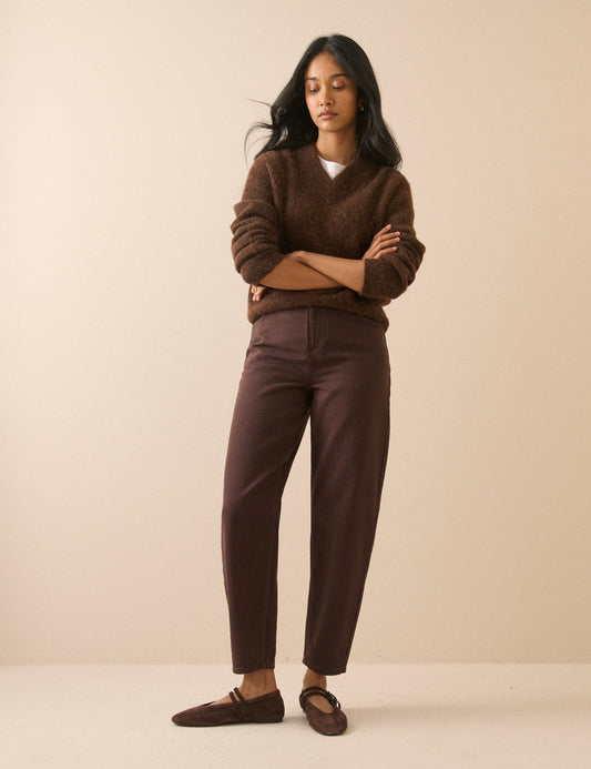 A woman stands with arms crossed wearing a brown fuzzy V-neck sweater, matching brown trousers, and brown slip-on shoes against a plain beige studio background.