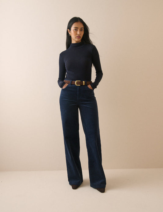 A woman stands against a plain beige studio background wearing a dark navy ribbed turtleneck top, dark blue wide-leg corduroy pants, and a brown leather belt with a gold buckle.