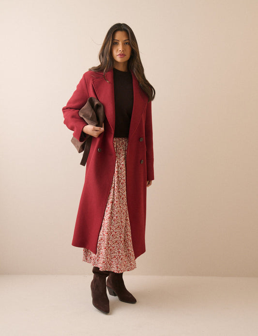 A woman stands against a plain beige studio background wearing a long burgundy coat over a black top and a pink floral midi skirt, with dark brown ankle boots and holding a dark brown jacket.