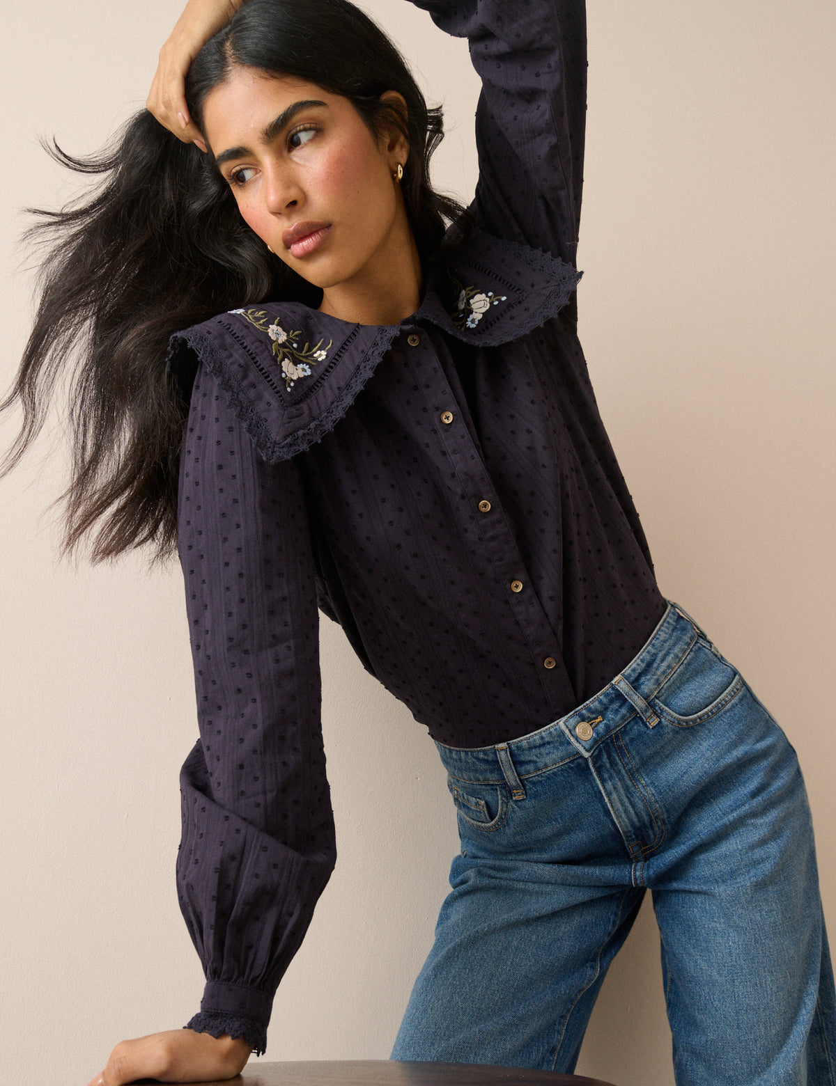 A woman wearing a dark navy long-sleeve blouse with a large ruffled collar decorated with small floral embroidery and buttons, paired with blue jeans, posing against a solid beige background.