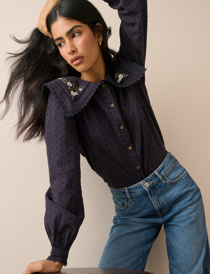 A woman wearing a dark navy long-sleeve blouse with a large ruffled collar decorated with small floral embroidery and buttons, paired with blue jeans, posing against a solid beige background.
