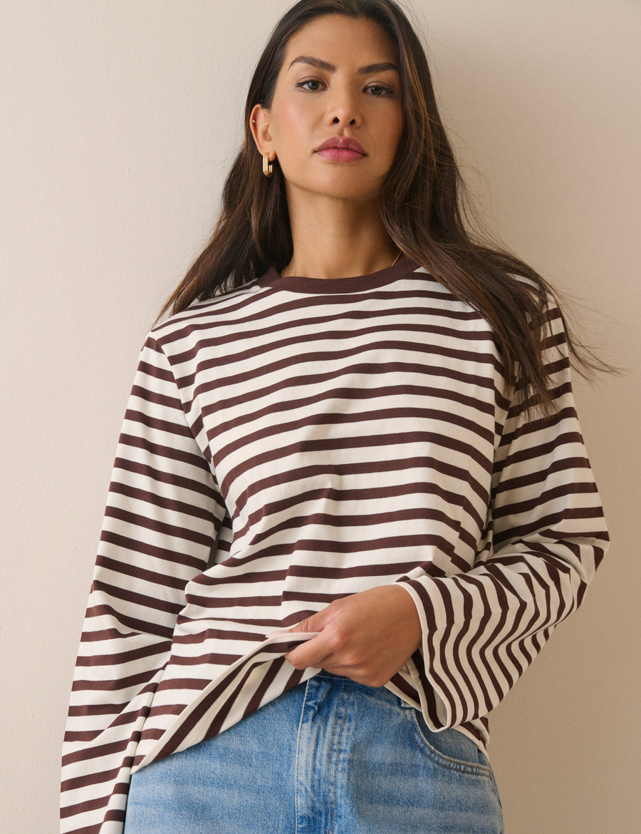 A woman wearing a brown and white horizontal striped long-sleeve top with blue jeans, standing against a plain beige background.