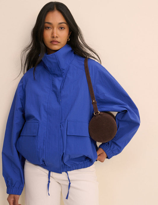 A woman wearing a bright royal blue lightweight jacket with a high collar, front zipper, and flap pockets, paired with white pants and a brown crossbody bag, set against a plain beige background.