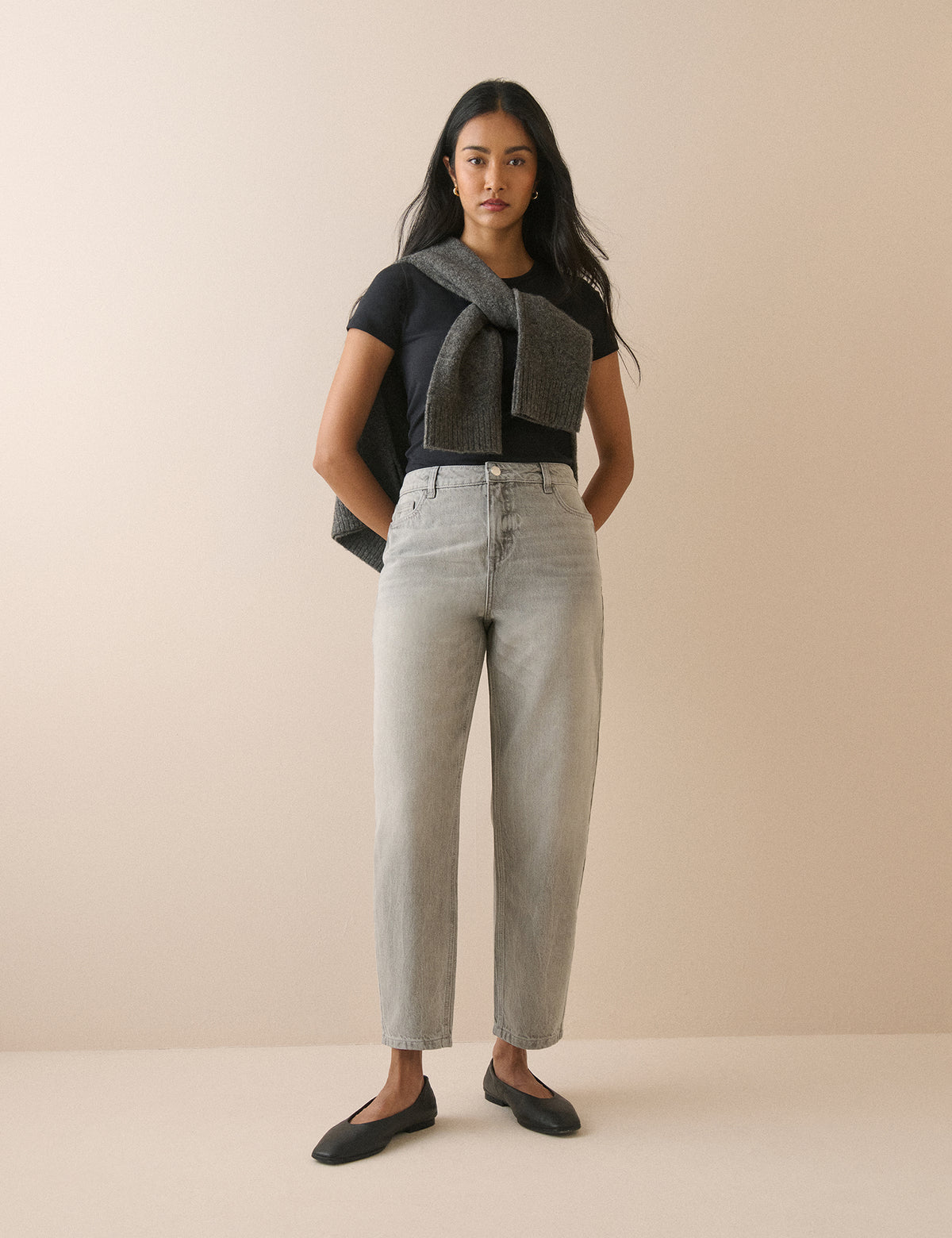 A woman stands against a plain beige studio background wearing a black short-sleeve top, a dark gray knitted scarf tied around her shoulders, light gray high-waisted straight-leg jeans, and black flats.