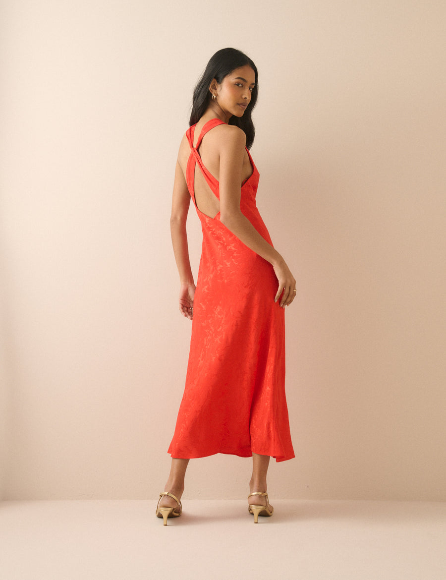 A woman modeling a bright coral-red sleeveless maxi dress with a textured lace overlay and a cutout back, standing against a solid beige studio backdrop.
