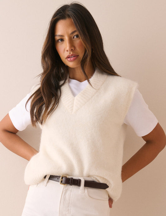 A woman wearing a cream-colored fuzzy knit sweater vest over a white t-shirt, paired with white pants and a dark brown belt, posing against a solid beige background.