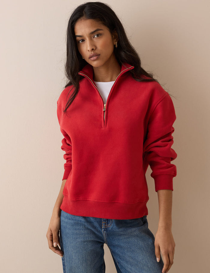 A woman wearing a bright red half-zip sweatshirt with a white undershirt and blue jeans, standing against a solid beige background.