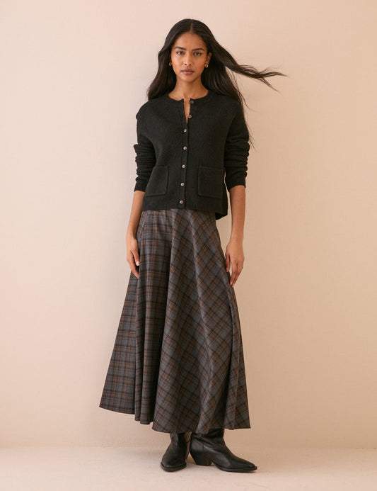 A woman stands against a plain beige studio background wearing a black cropped button-front cardigan, a long grey and brown tartan skirt, and black ankle boots.