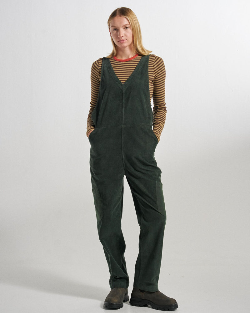 A woman modeling dark green corduroy dungarees over a brown and beige striped long-sleeve shirt, standing against a light grey studio background.