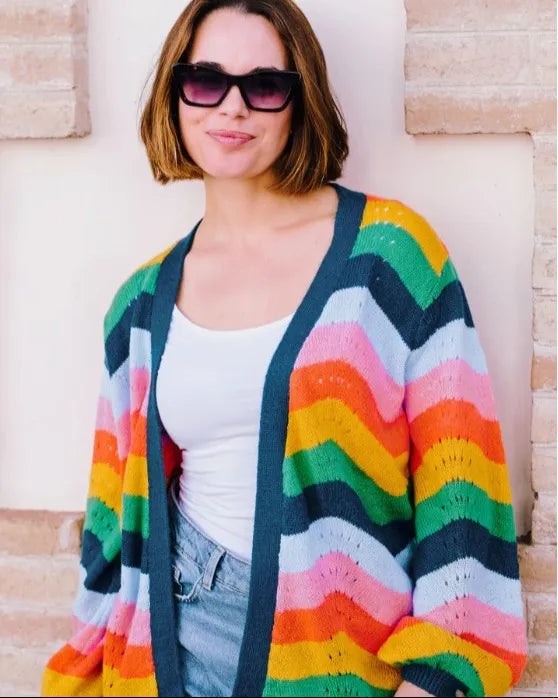 Woman wearing a striped cardigan in Spring rainbow colours, with a white top and jeans, standing by a stone wall.