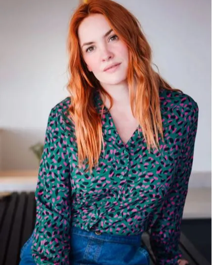 Woman with red hair wearing a patterned blouse of deep green with black and pink flashes, against a neutral background.