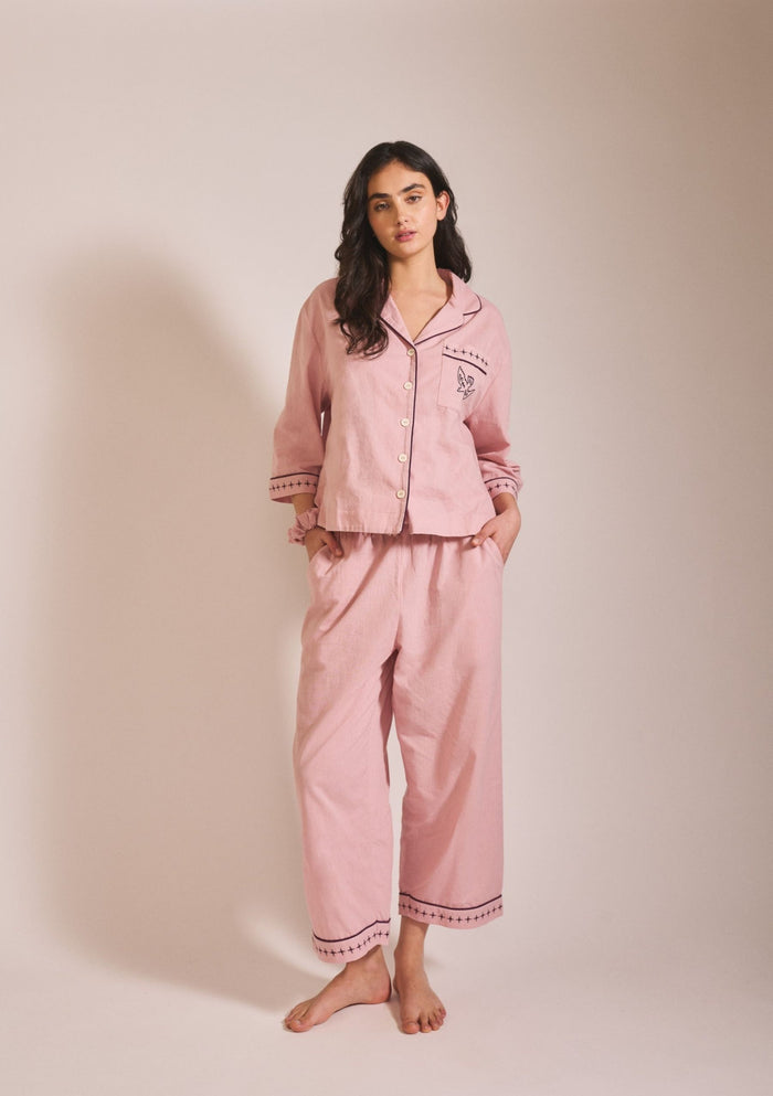 Model wearing a relaxed-fit pink pyjama set in a soft, muted dusty rose tone, with long sleeves, button front, and loose trousers.