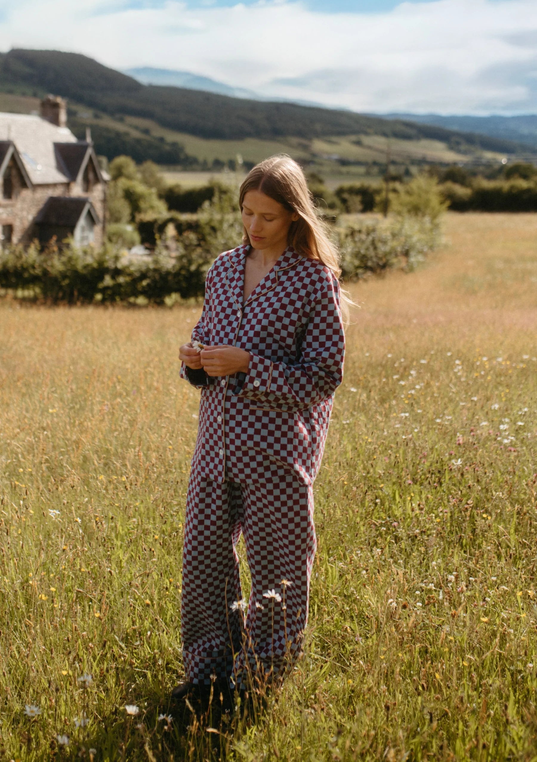 Woman in a checkered pyjama outfit standing in a field with a house and mountains in the background. Pyjamas are by TBCo, are burgundy and blue, and  filtered by Shop My Palette for the Cool Summer and Light Summer colour seasons.