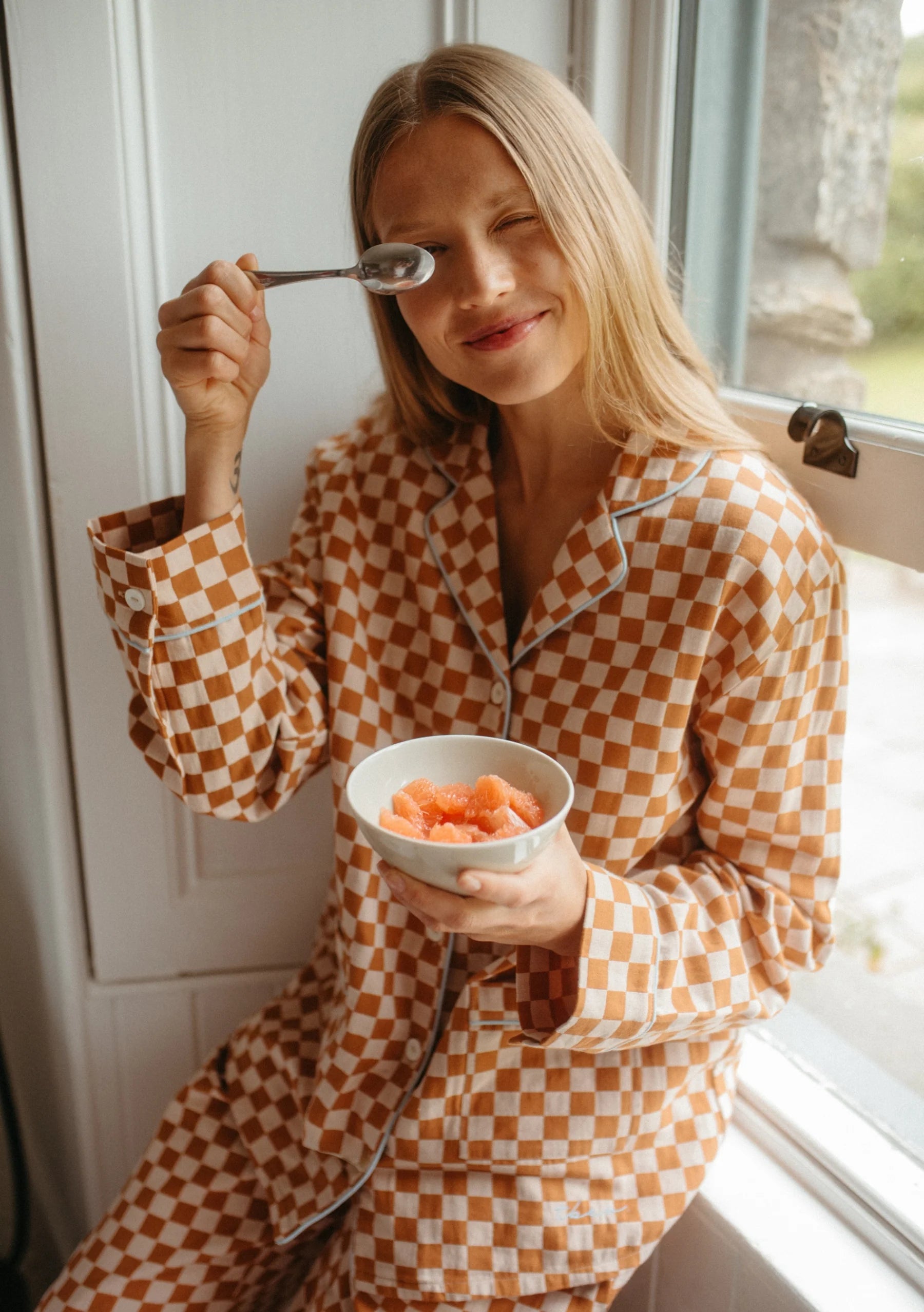 Woman in checkered pyjamas eating from a bowl by a window. Pyjamas are by TBCo and are filtered by Shop My Palette for the Light Spring, Warm Spring, Warm Autumn, and Soft Autumn colour seasons.