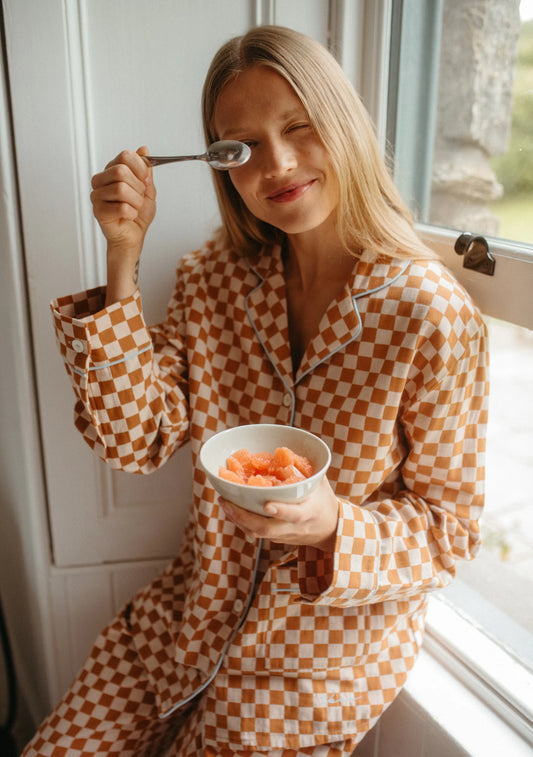 Woman in checkered pyjamas eating from a bowl by a window. Pyjamas are by TBCo and are filtered by Shop My Palette for the Light Spring, Warm Spring, Warm Autumn, and Soft Autumn colour seasons.
