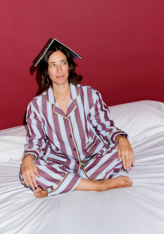 Model sits on a bed with a book on her head, wearing light blue and maroon striped pyjamas. Pyjamas are by TBCo x Glassette and are filtered by Shop My Palette for the Cool Summer and Light Summer colour seasons.