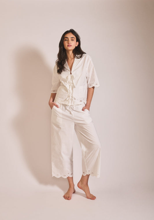 Model wearing a relaxed off-white pyjama set with short sleeves, tie-front detail, and wide-leg cropped trousers with delicate broderie trim.