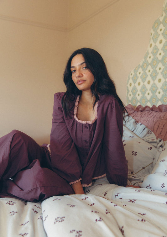 A woman sits on a bed wearing a matching mauve-purple pajama set with light pink lace trim. The bedroom has beige walls, patterned pillows, and a decorative headboard.