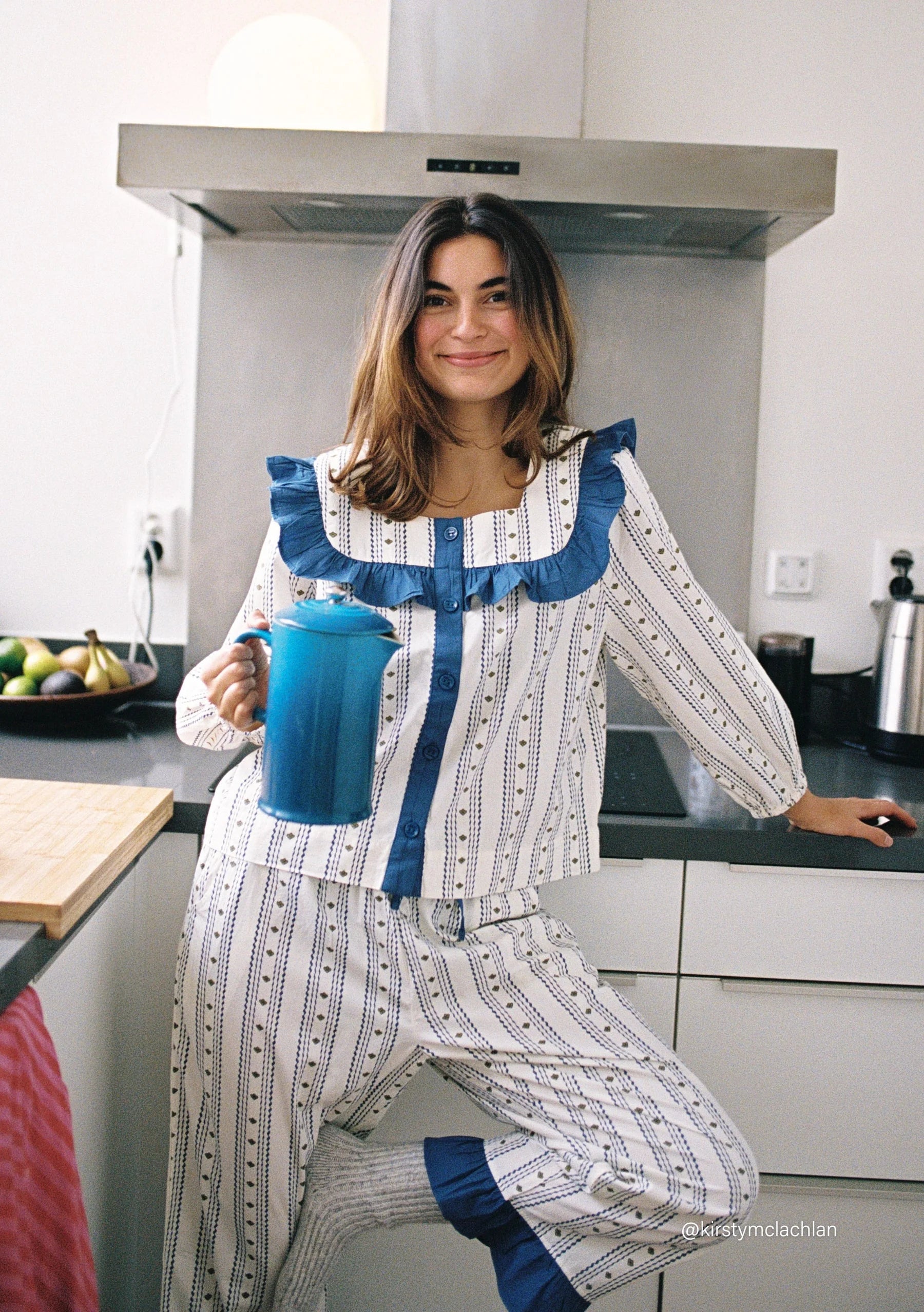 Woman in a blue and white patterned pyjama set, holding a blue cafetiere in a kitchen. Pyjamas are by TBCo and are filtered by Shop My Palette for the Cool Summer and Light Summer colour seasons.