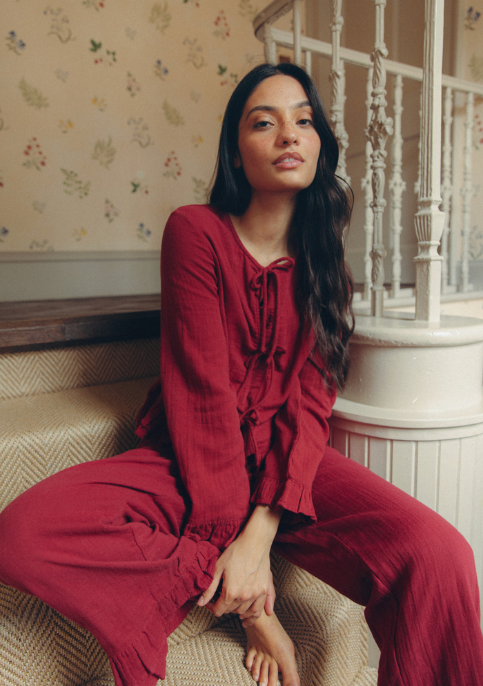 A woman wearing a matching red long-sleeve top and wide-leg trousers set sits on a beige herringbone staircase in a home interior with floral wallpaper and a white railing.