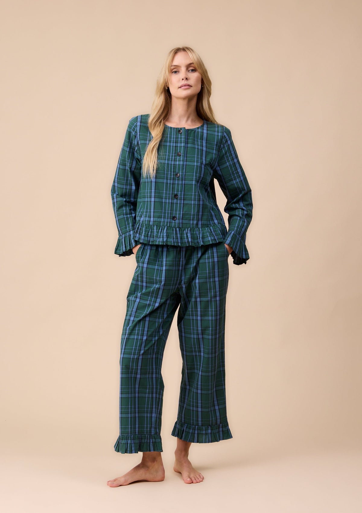 A woman modelling a two-piece tartan pyjama set consisting of a long-sleeve button-front top and matching pants, in a dark green and blue tartan pattern, against a solid beige studio background.