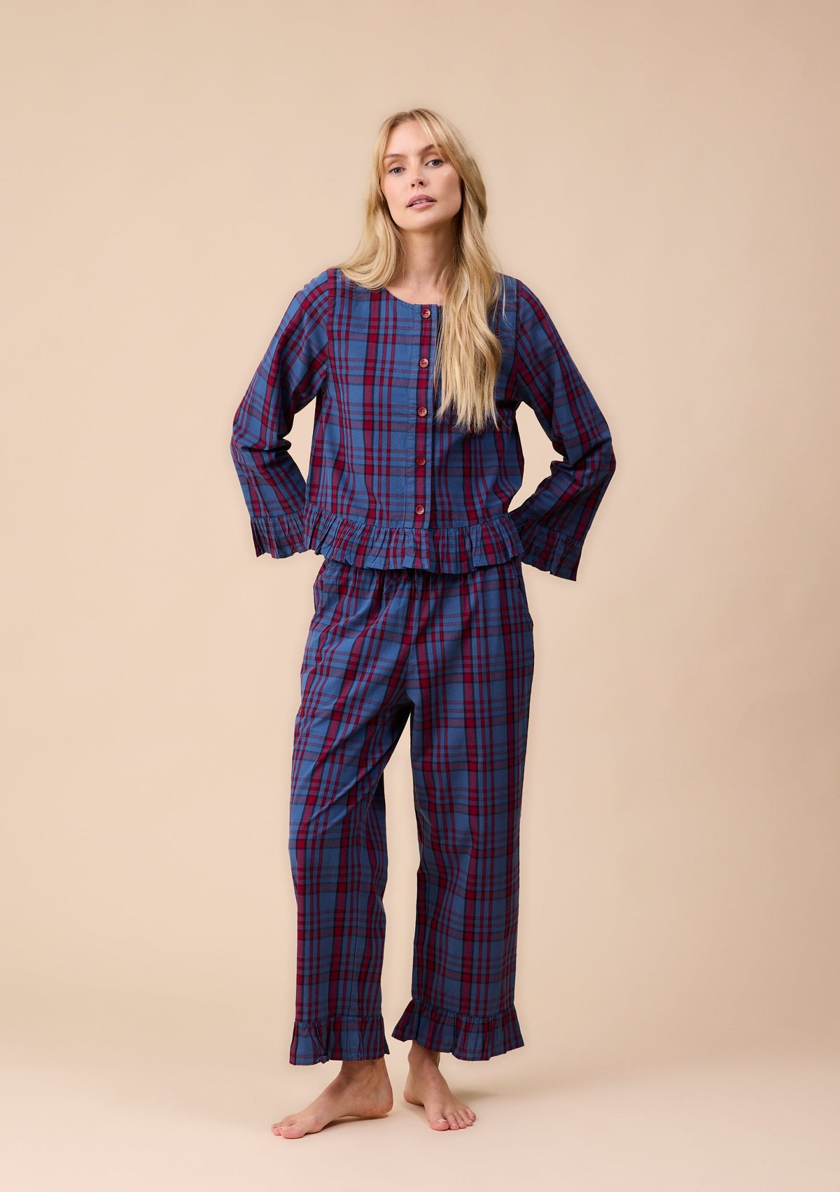 A woman modeling a two-piece plaid pyjama set consisting of a long-sleeve button-front top and matching pants, in a navy blue and red tartan pattern, against a solid beige studio background.