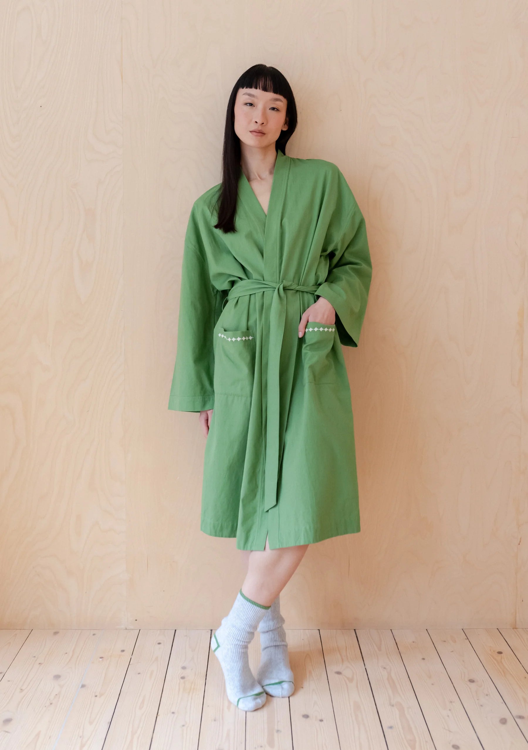 Model wearing a grass green cotton robe, leaning against a wooden wall. Robe is by TBCo and is filtered for the Clear Spring, Light Spring, and Warm Spring colour seasons by Shop My Palette.