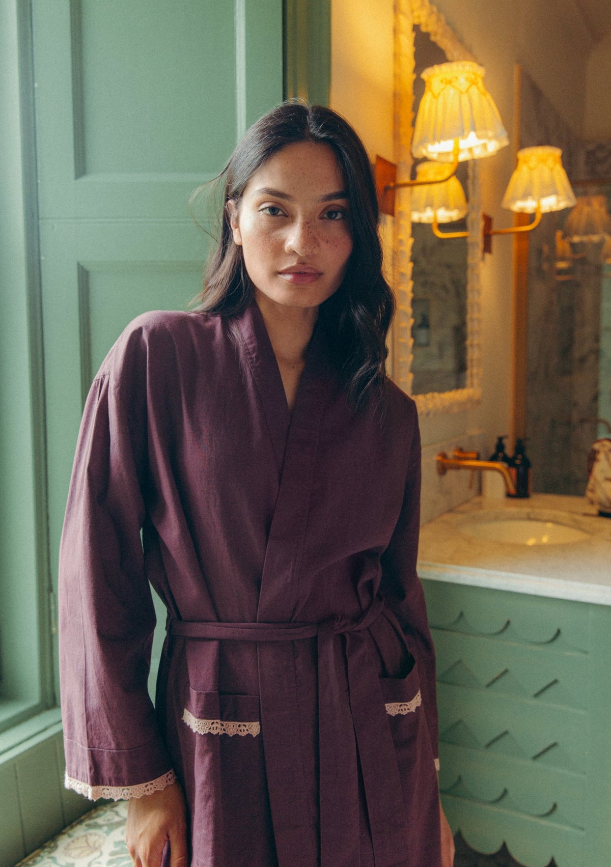 Model wearing a soft plum cotton robe with lace trim, standing in a bathroom with green wooden walls.