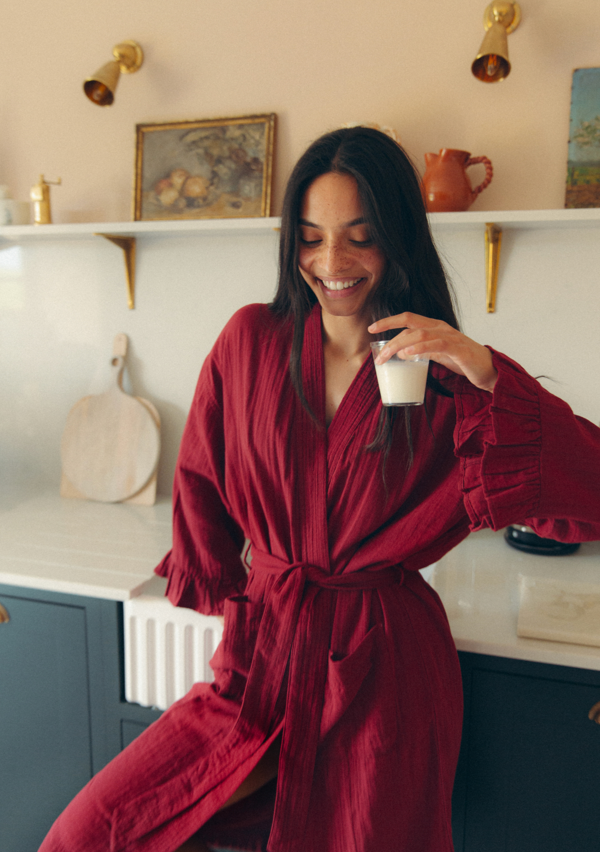 A woman wearing a textured, deep red wrap robe sits in a kitchen setting, holding a white mug. The background features white shelves with decorative items, brass wall sconces, and dark blue cabinets.