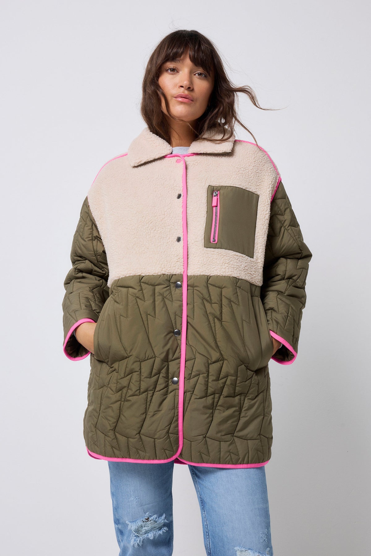 A model wearing a color-blocked quilted jacket with olive green, beige sherpa, and bright pink trim, paired with blue jeans, against a light gray studio background.