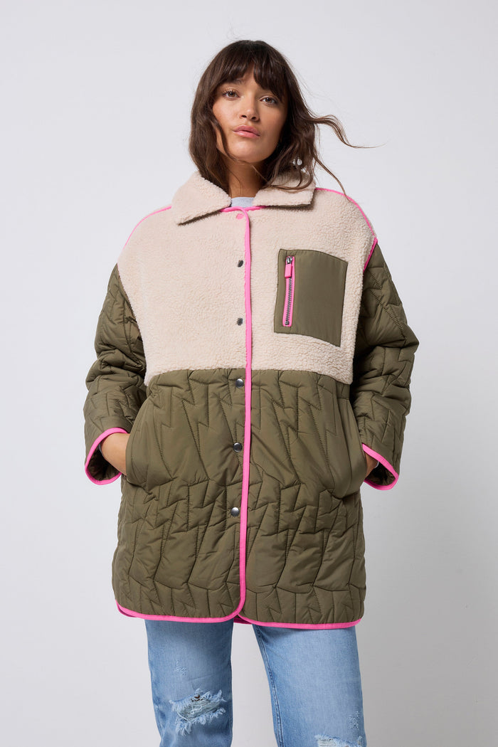 A model wearing a color-blocked quilted jacket with olive green, beige sherpa, and bright pink trim, paired with blue jeans, against a light gray studio background.