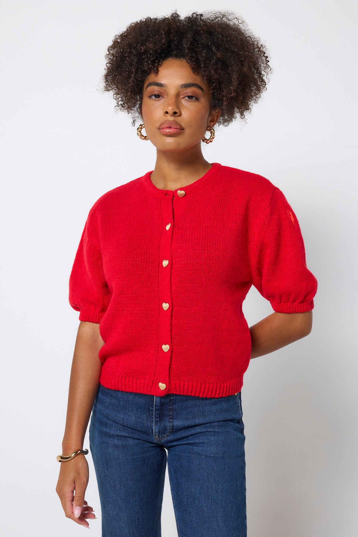 Model wears a bright red short sleeve cardigan with gold buttons, gold earrings, and denim jeans.