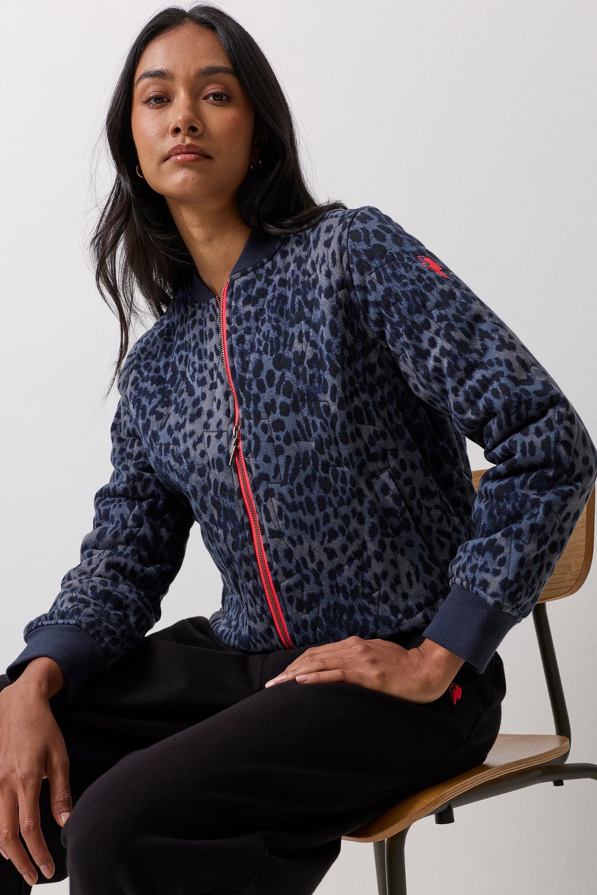 A woman wearing a navy blue leopard-print bomber jacket with a red zipper, seated on a wooden chair against a light gray background.