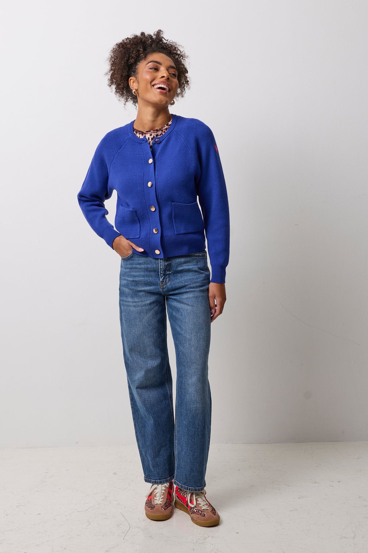 A woman modeling a vibrant royal blue knit cardigan with gold buttons, paired with blue jeans and patterned shoes, standing against a plain light gray studio background.