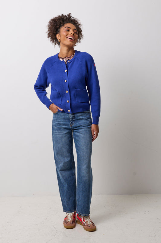 A woman modeling a vibrant royal blue knit cardigan with gold buttons, paired with blue jeans and patterned shoes, standing against a plain light gray studio background.