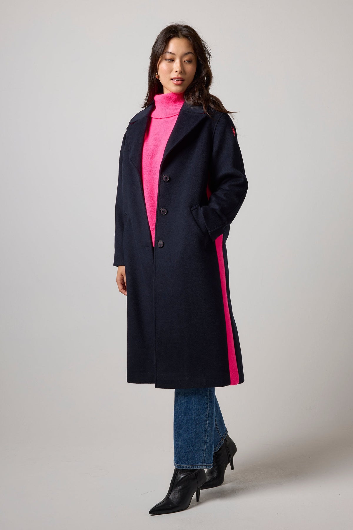 A woman models a long navy coat with bright pink side stripes, worn over a hot pink turtleneck sweater, blue jeans, and black ankle boots, against a neutral gray studio background.