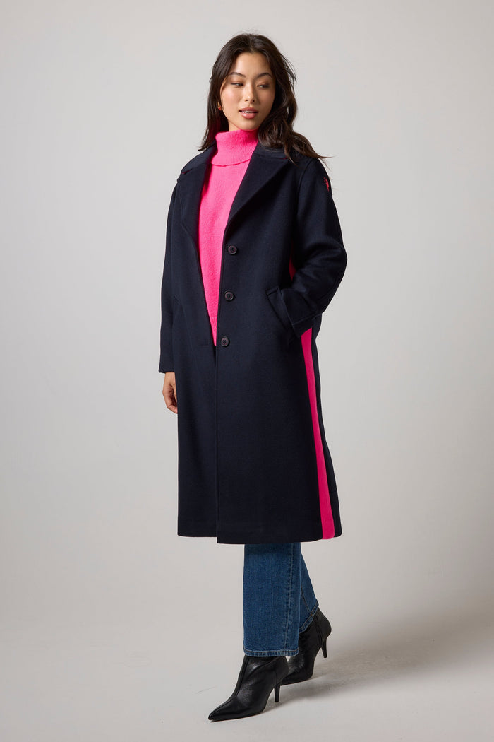 A woman models a long navy coat with bright pink side stripes, worn over a hot pink turtleneck sweater, blue jeans, and black ankle boots, against a neutral gray studio background.