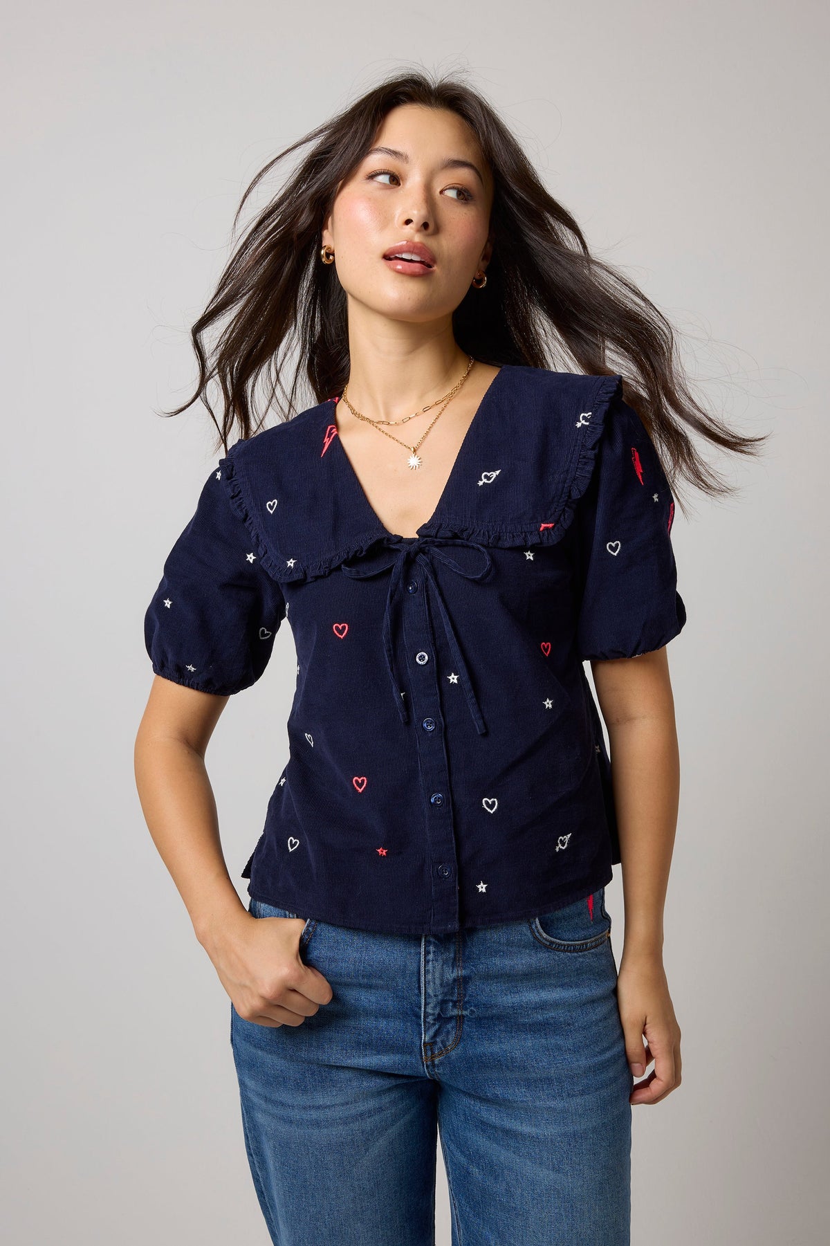 A woman modeling a navy blue short-sleeve top with embroidered white and red hearts, paired with blue jeans, against a light gray studio background.