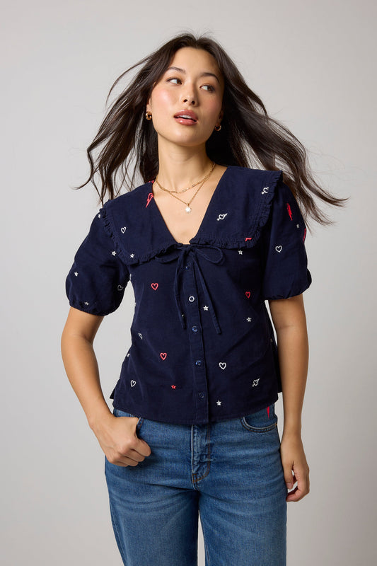 A woman modeling a navy blue short-sleeve top with embroidered white and red hearts, paired with blue jeans, against a light gray studio background.