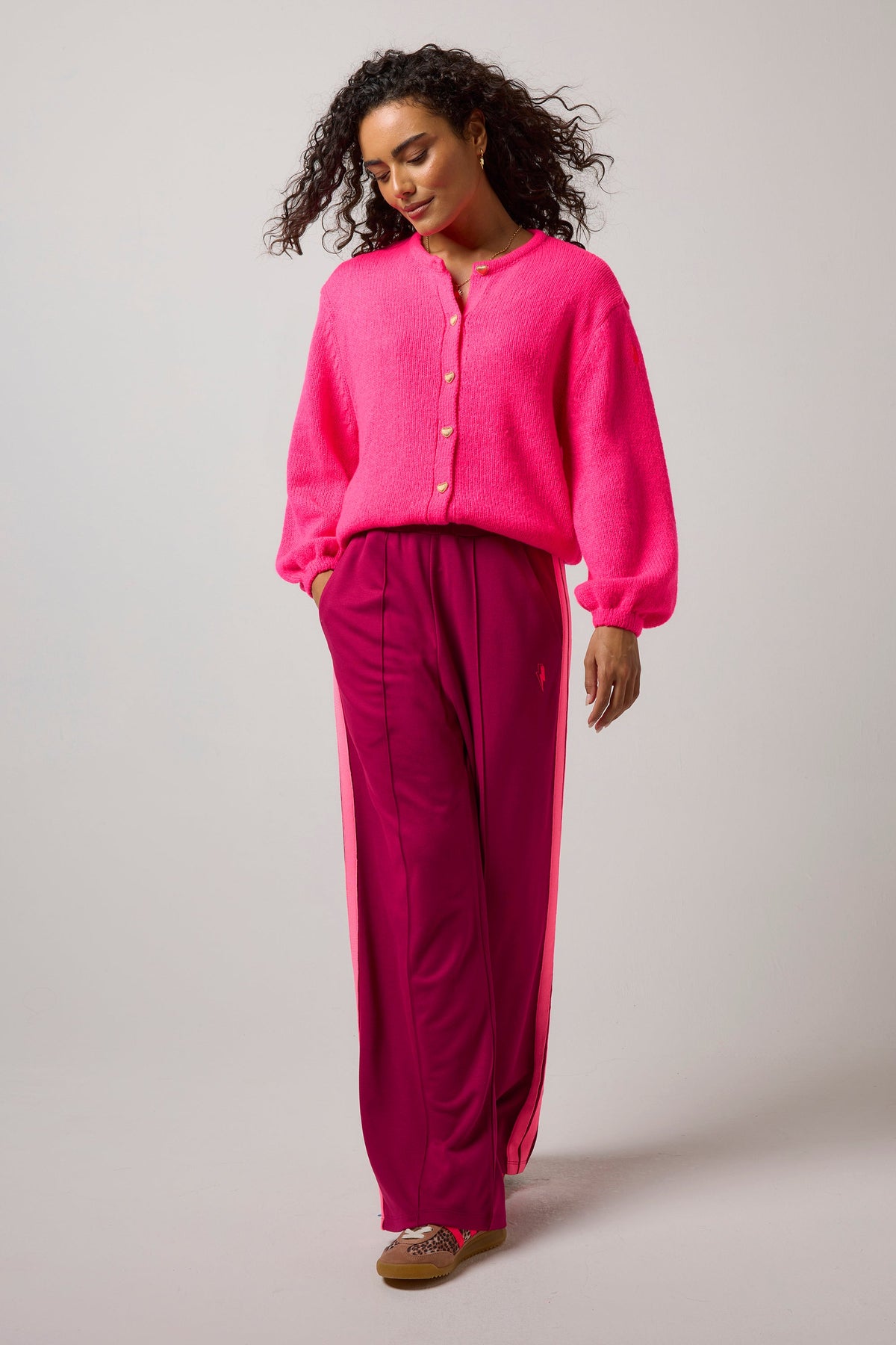 A woman modeling a bright pink fuzzy long-sleeve button-front top and magenta wide-leg pants with pink side panels, standing against a light gray studio background.