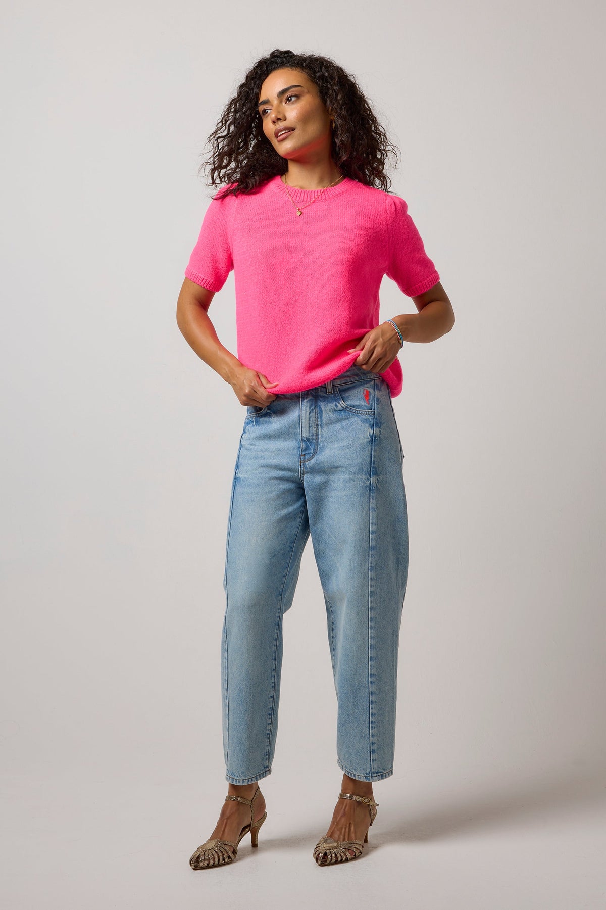 A woman modeling a bright pink short-sleeve knit top and light-wash blue jeans against a plain light gray studio background.