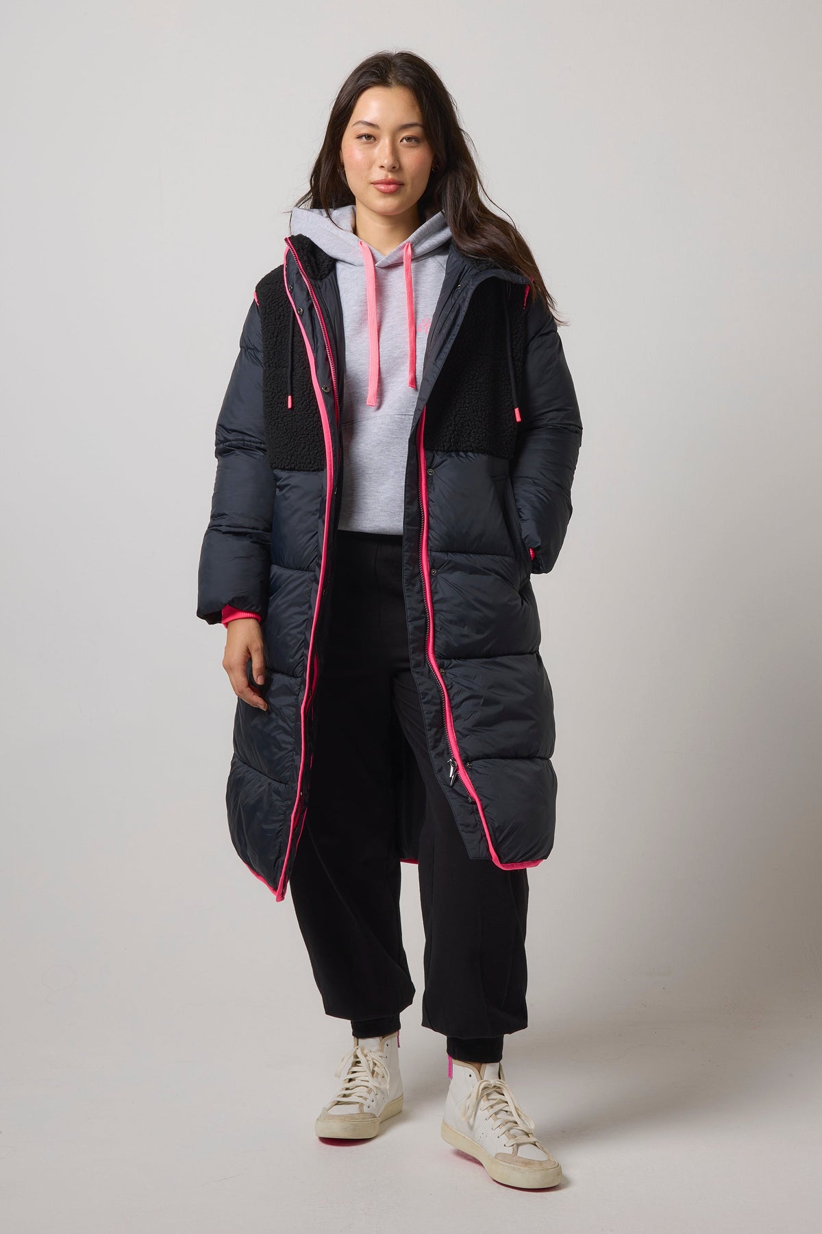 A woman modeling a long black puffer coat with bright pink piping and a pink-lined hood, worn over a light gray hoodie, black sweatpants, and off-white sneakers against a light gray studio background.
